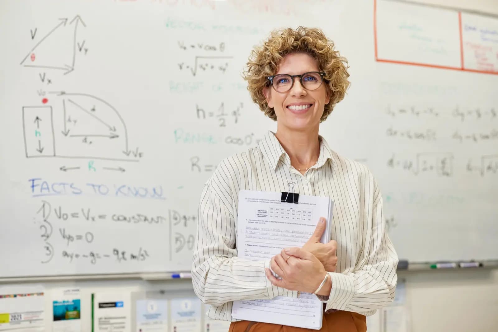 Teacher smiling in front of a whiteboard, holding a student's homework